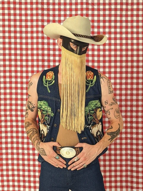 Person wearing cowboy hat, black mask with fringe, and embroidered denim vest against a red and white checkered backdrop.