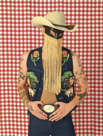 Person wearing cowboy hat, black mask with fringe, and embroidered denim vest against a red and white checkered backdrop.