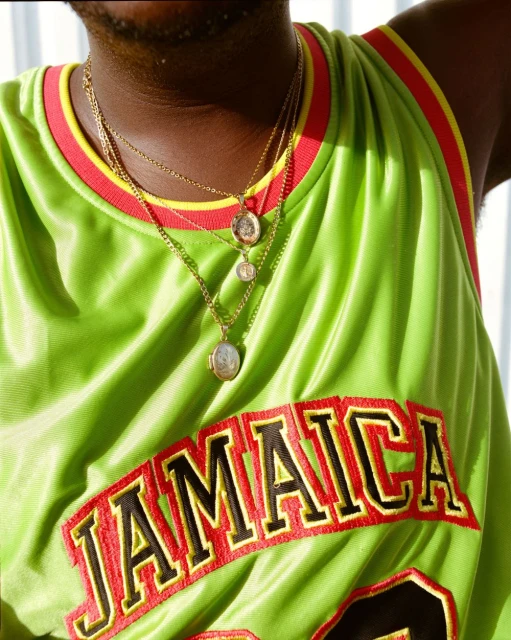 Close-up of a dark-skinned man in a green jersey with 'JAMAICA' on it, wearing layered gold chains and pendants against a white background.