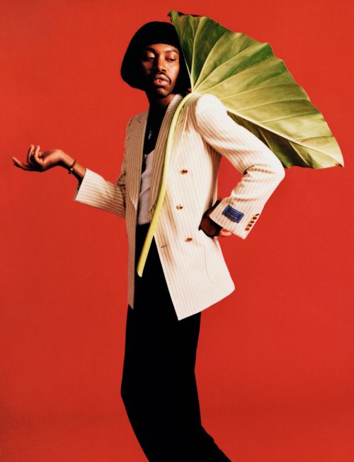 A stylish Black man in a white suit and black pants poses against a red backdrop, holding a large tropical leaf over his shoulder.