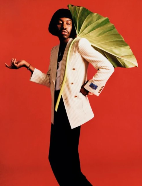 A stylish Black man in a white suit and black pants poses against a red backdrop, holding a large tropical leaf over his shoulder.