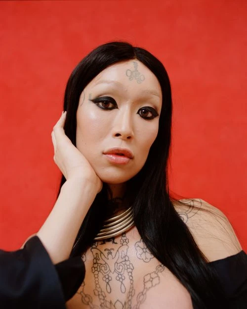 Portrait of a person with black hair and face tattoos against a red background, wearing a gold necklace and black clothing.