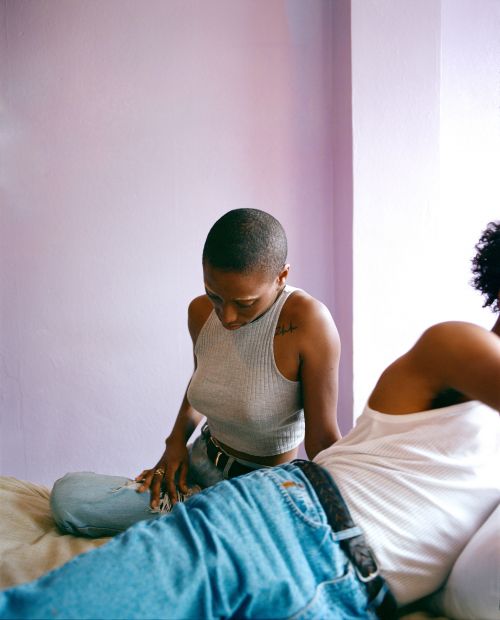 Two dark-skinned women sit on a bed in a room with lavender walls and natural light, both wearing casual tops and blue jeans.