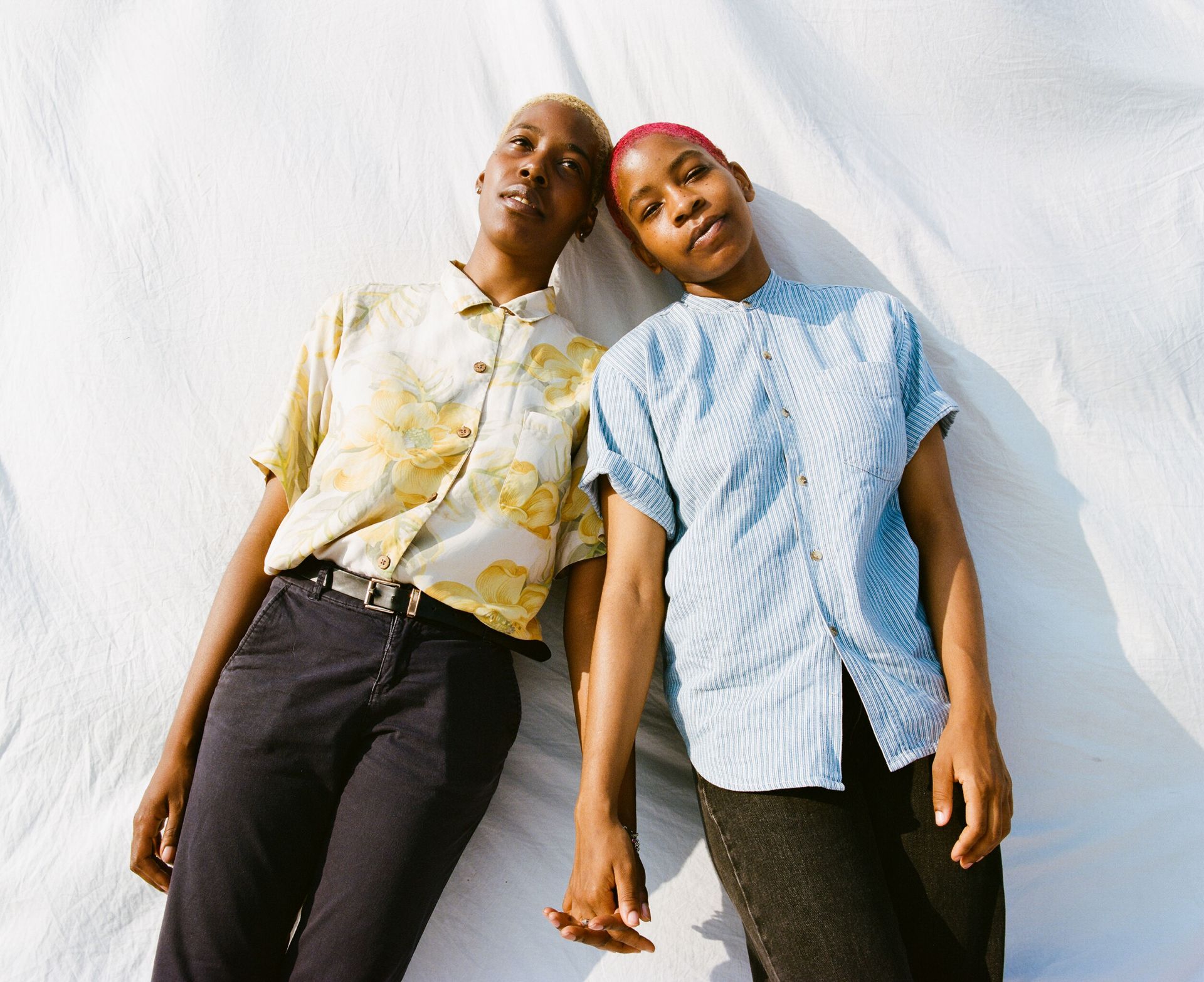 Two women with short hair pose together in front of a white backdrop, wearing casual shirts and dark pants, holding hands in a studio-like setting.