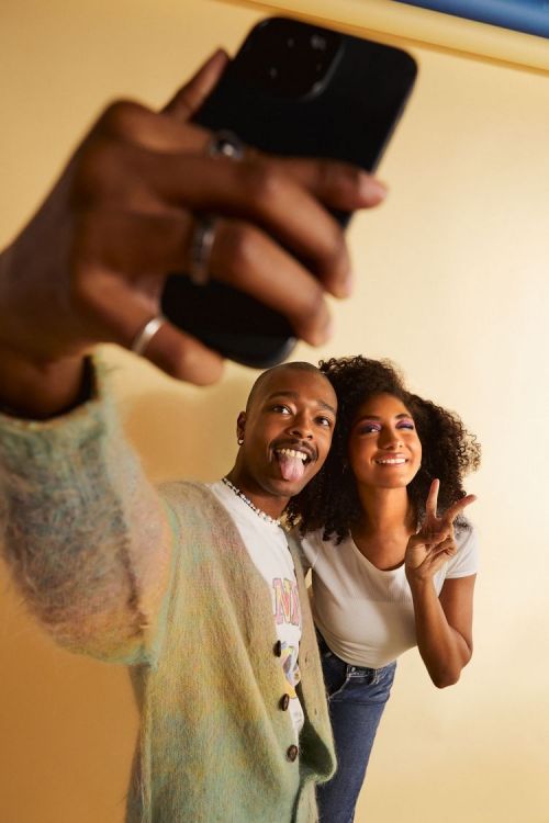 Two adults smiling and posing for a selfie using a smartphone, wearing casual outfits indoors with a bright studio lighting setup.