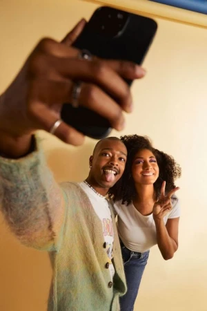 Two adults smiling and posing for a selfie using a smartphone, wearing casual outfits indoors with a bright studio lighting setup.
