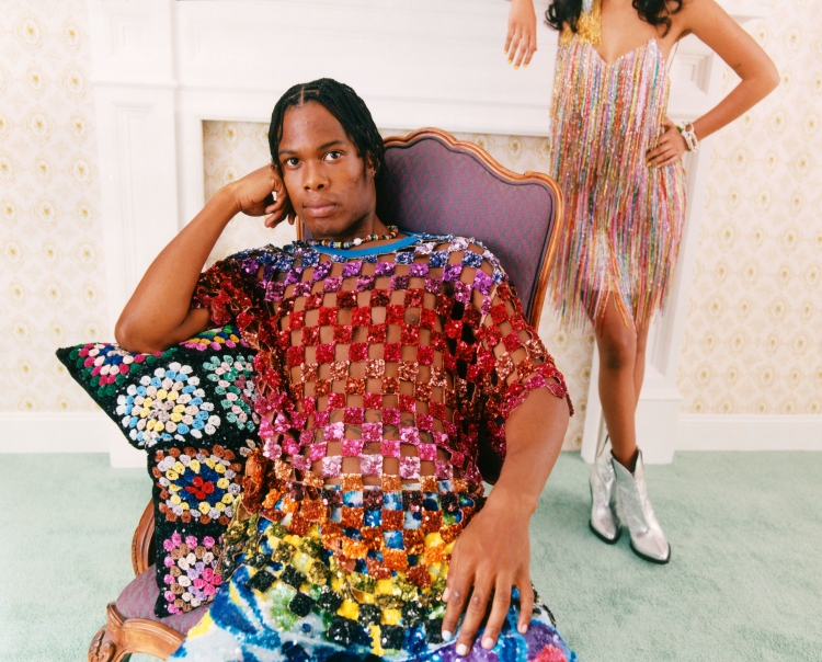 An adult male sits in a chair wearing a multicolored sequined shirt, a woman in a sequined dress and silver cowboy boots stands behind him.