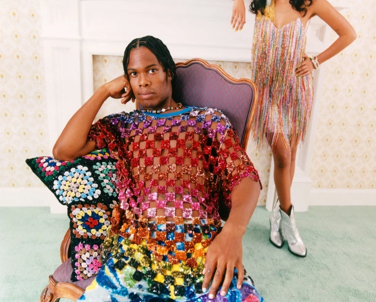An adult male sits in a chair wearing a multicolored sequined shirt, a woman in a sequined dress and silver cowboy boots stands behind him.