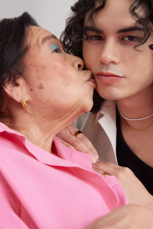 Elderly woman in pink shirt kissing a young adult with curly hair in a studio setting. They both wear gold jewelry.