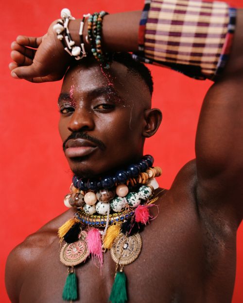 Close-up portrait of a Black male model with glitter makeup, wearing beaded jewelry and arm raised against a red studio backdrop.