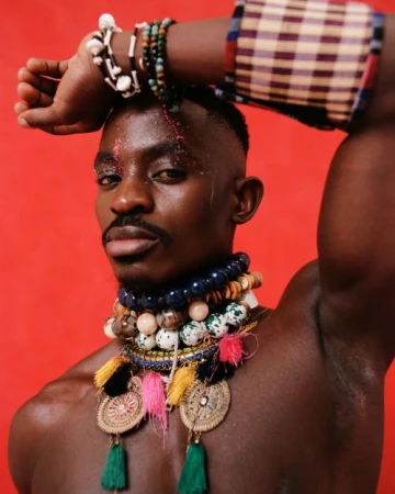 Close-up portrait of a Black male model with glitter makeup, wearing beaded jewelry and arm raised against a red studio backdrop.