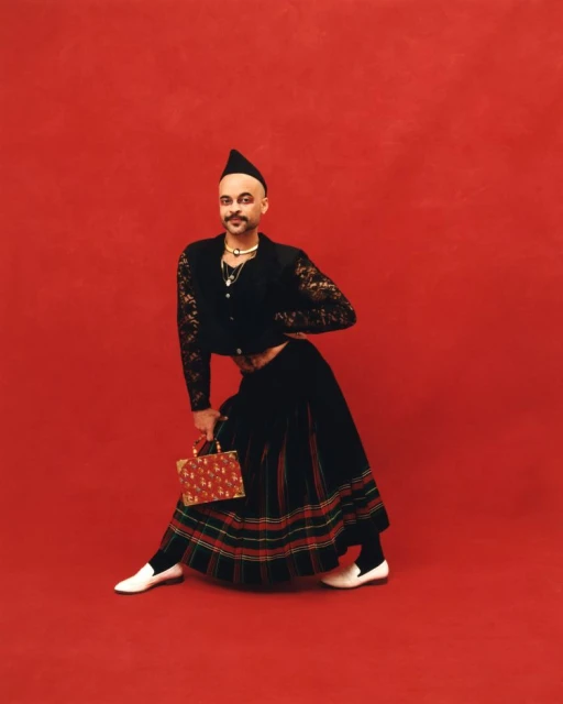 A stylish adult male poses against a vibrant red backdrop wearing a black lace top, kilt, and holding a red purse.