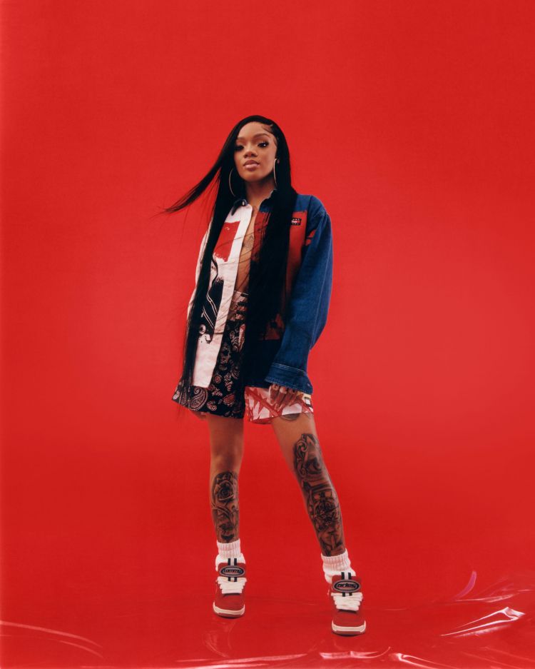 A young woman with long dark hair poses in a patterned outfit and platform shoes against a vibrant red backdrop in a studio shot.