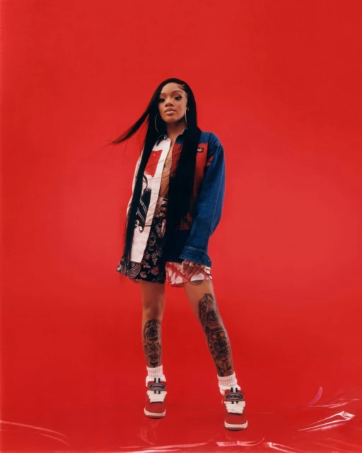 A young woman with long dark hair poses in a patterned outfit and platform shoes against a vibrant red backdrop in a studio shot.