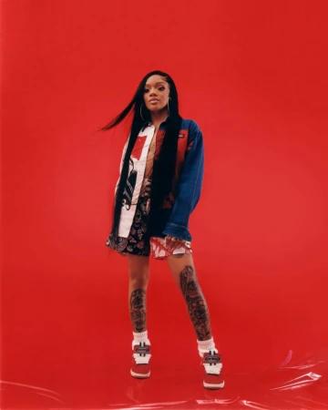 A young woman with long dark hair poses in a patterned outfit and platform shoes against a vibrant red backdrop in a studio shot.
