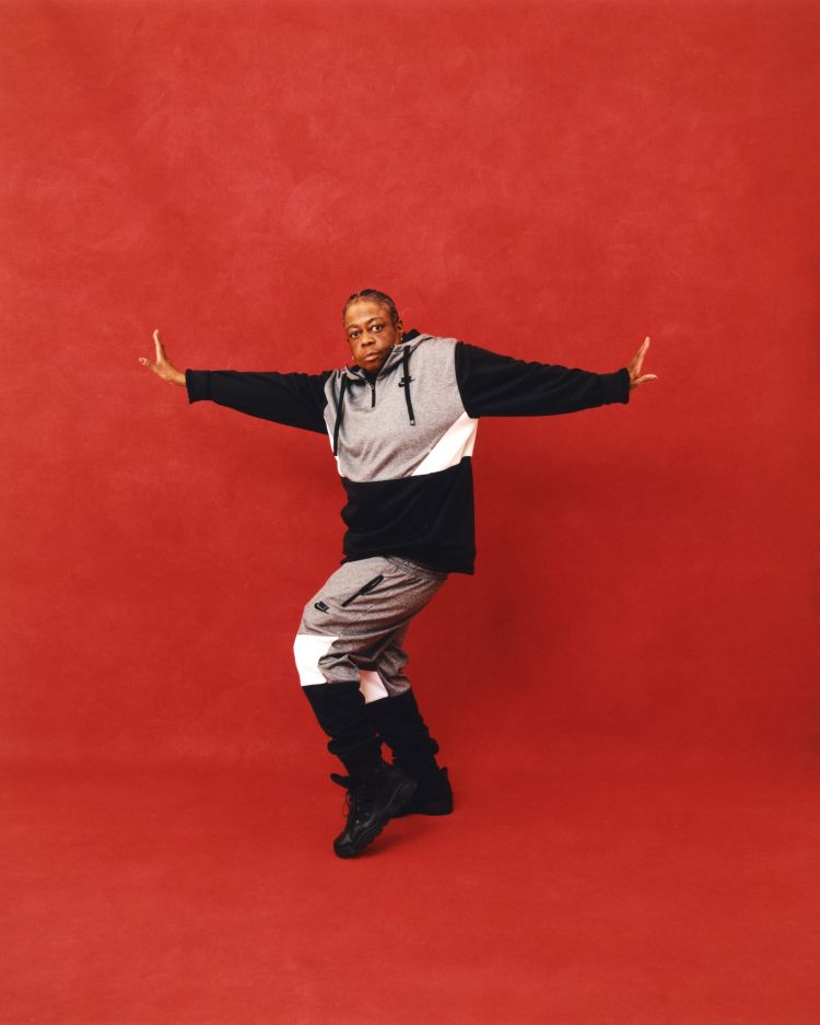 Black man wearing a color-blocked hoodie and sweatpants posing against a red textured backdrop