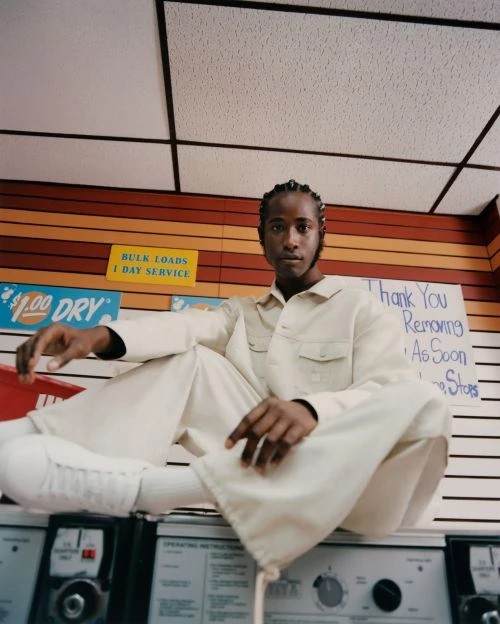 Adult male sitting cross-legged on top of a row of washing machines in a laundromat, wearing a tan, long-sleeved jumpsuit.