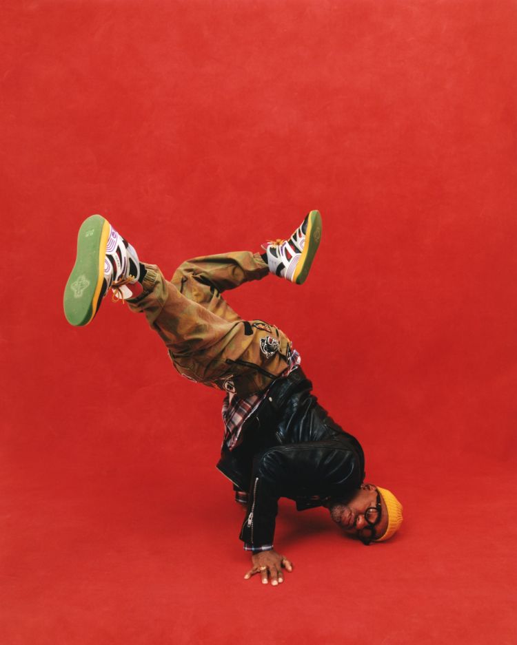 Person does a handstand on red background wearing yellow beanie, glasses, black jacket, plaid shirt, and tan pants with sneakers.