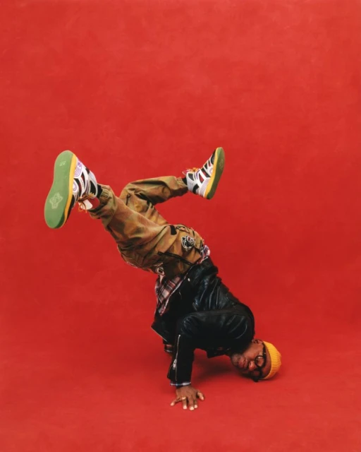 Person does a handstand on red background wearing yellow beanie, glasses, black jacket, plaid shirt, and tan pants with sneakers.