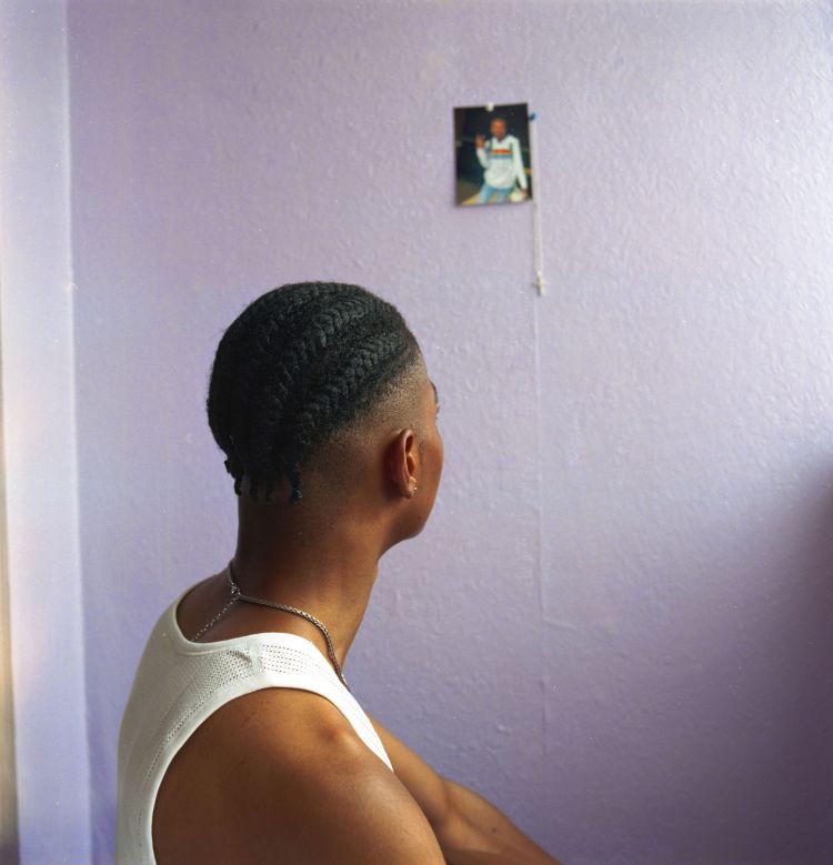 Young adult man with braided hair looking at a photograph on a lavender wall, wearing a white tank top and silver necklace indoors.