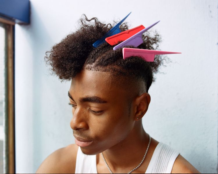 Young adult with dark hair in a tank top poses indoors, multiple combs stuck in his afro, lit by natural light from a nearby window.