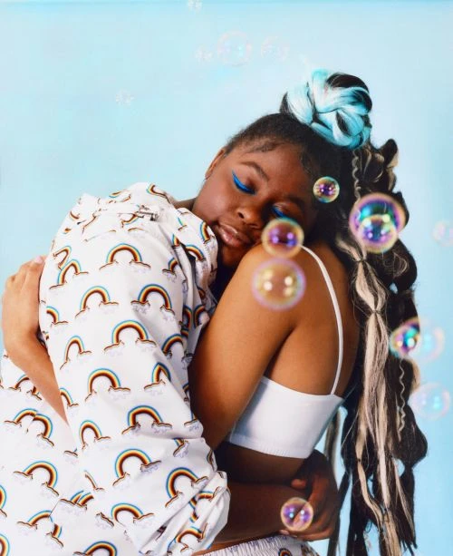Two women hug against a light blue background with floating soap bubbles, one wearing rainbow print pajamas and one wearing a white tank top.