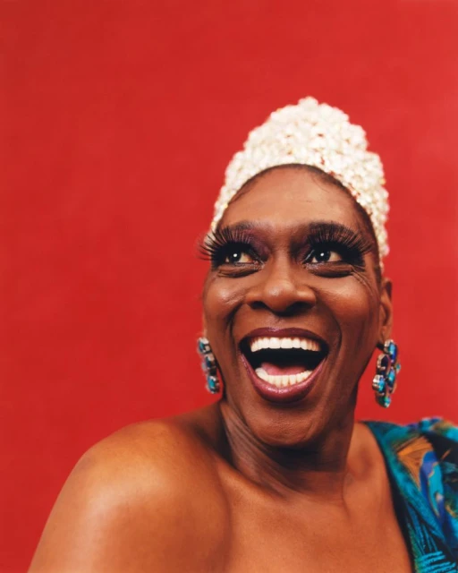 Portrait of a smiling woman with dark skin, large eyelashes, beaded cap, and colorful shoulder wrap against a red background.