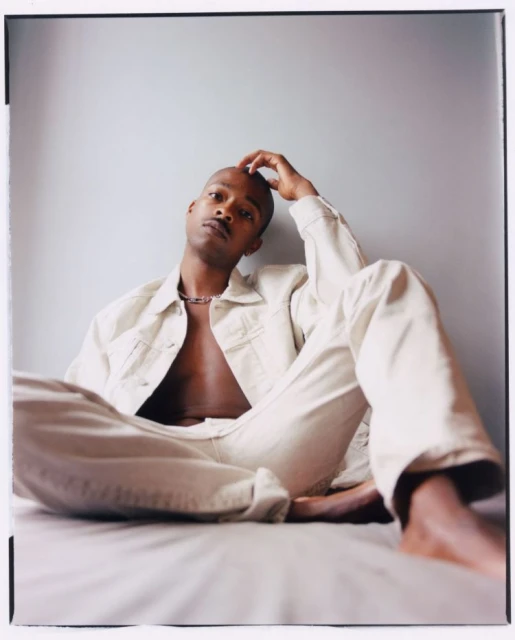 Portrait of an African American male sitting cross-legged on a bed, wearing a cream-colored denim jacket and matching pants, casually posed indoors.
