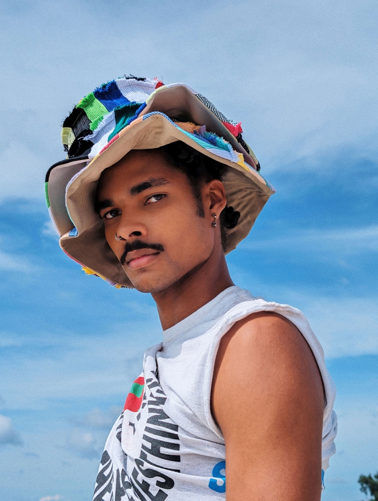Portrait of a young adult Black male with a mustache and a colorful patchwork bucket hat posing outdoors against a blue sky backdrop.