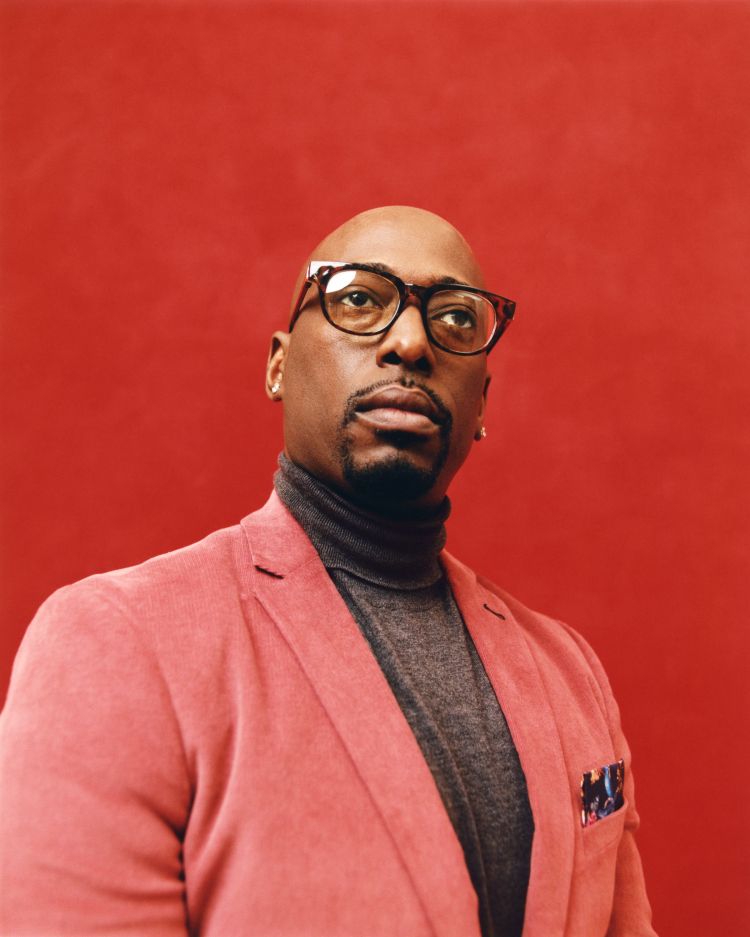 Portrait of a bald Black man with glasses, wearing a grey turtleneck sweater and a red corduroy blazer against a red background.
