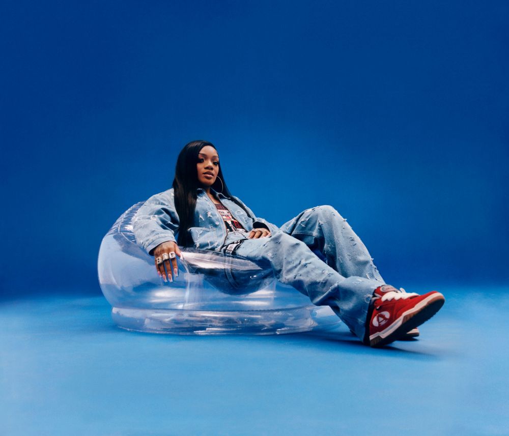 Stylish adult woman seated on clear inflatable chair against vibrant blue backdrop, wearing denim outfit and red sneakers, confident gaze.