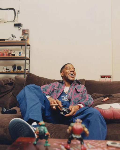 A man relaxing on a brown couch in a casual pose, engrossed in a video game with Teenage Mutant Ninja Turtle figures on a table.