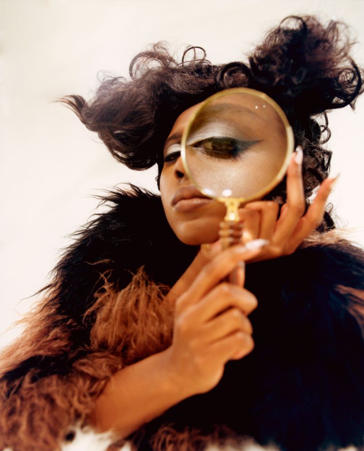 Close-up of an African American woman with an afro hairstyle, wearing a fur-trimmed jacket and holding a magnifying glass to her eye.