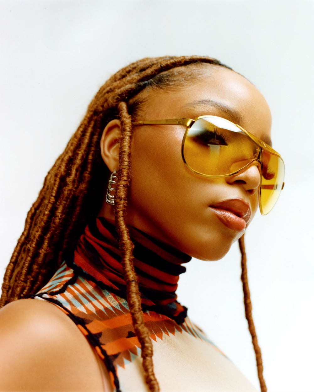 A medium shot captures a woman with auburn dreadlocks, statement gold sunglasses, and a sleeveless geometric print top against a white backdrop.
