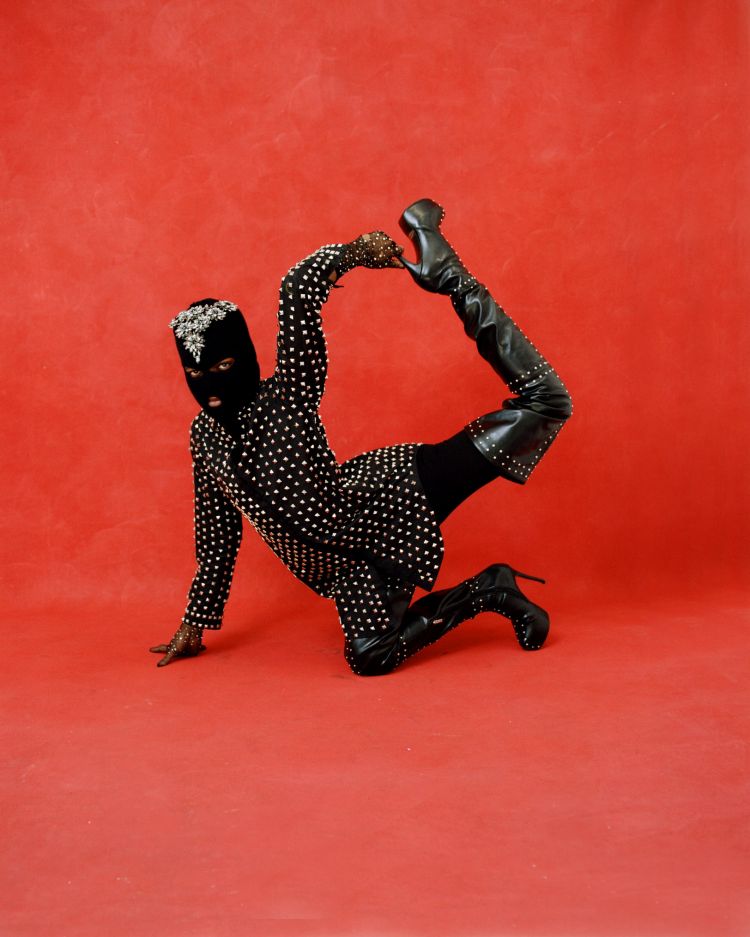 Person in black studded jumpsuit and high-heeled boots poses on a red seamless background, showcasing a dramatic, stylized fashion statement.