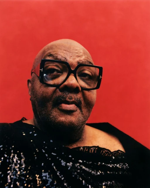 Portrait of a Black adult with black glasses and a sequin top against a red background in a studio setting with neutral lighting.