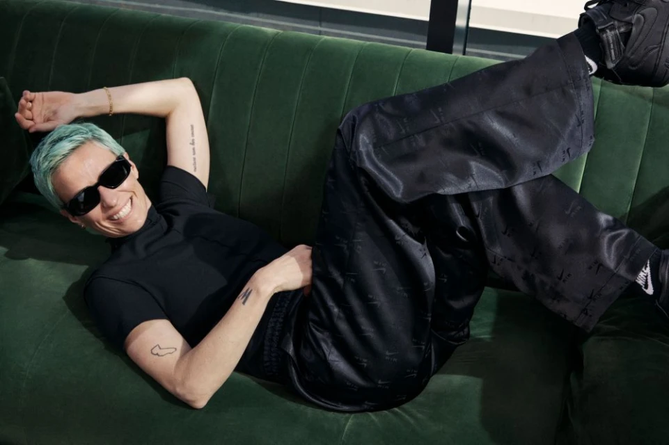Woman with blue hair and sunglasses reclines on a green velvet couch wearing black top and pants, black sneakers, and smiling indoors.
