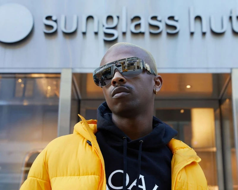 Man with sunglasses in an urban scene, wearing a yellow puffer jacket and black hoodie, standing in front of a Sunglasses Hut store.