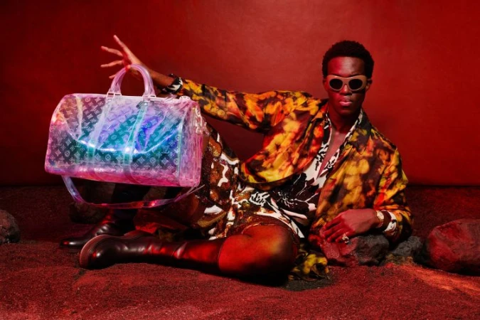 Black man holding a translucent monogram duffel bag while wearing patterned clothing and sunglasses on a red set.