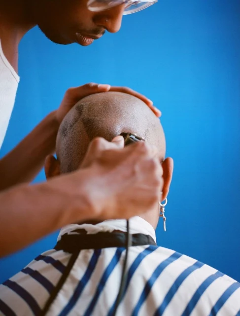 Man getting a haircut by barber wearing safety glasses in a blue photo studio setting. Professional grooming service with electric razor.
