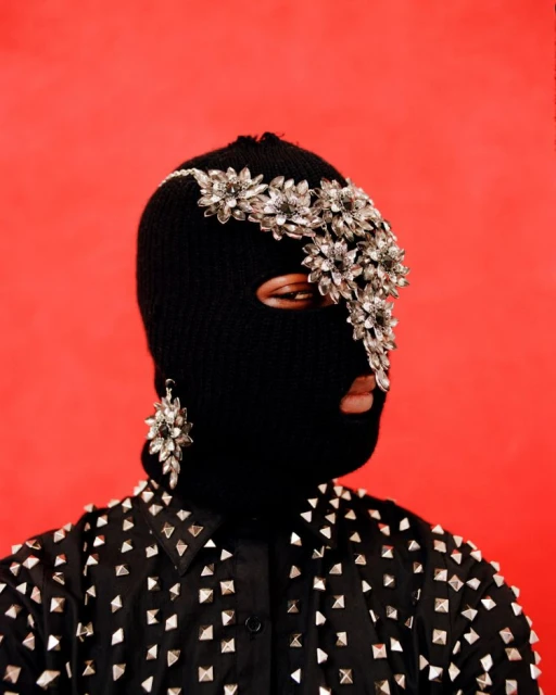 Person wearing a black balaclava and embellished headpiece, earring and studded black top against a solid red background.