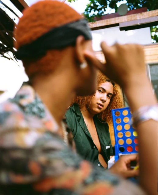 Two women play a board game outdoors, one in focus and one blurred in the foreground, in casual clothing under natural light.