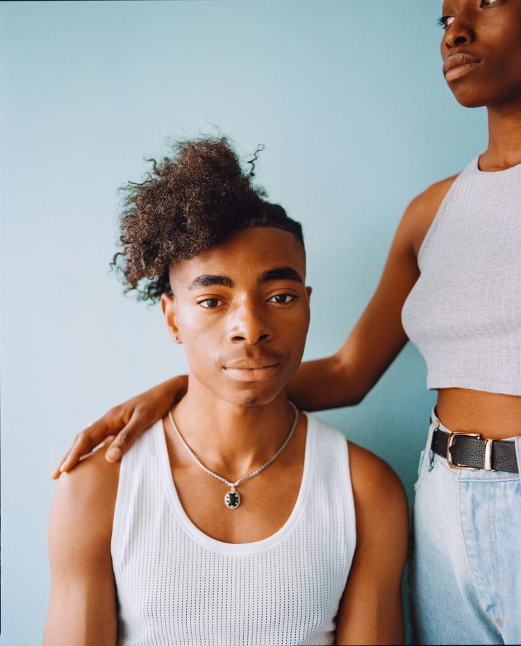 Portrait of a young Black man in a white tank top with a woman behind him placing her hand on his shoulder against a blue background.