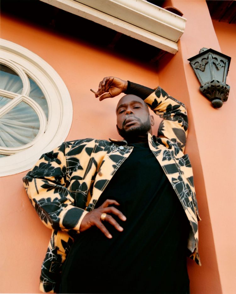 Stylish man in patterned jacket leans against peach wall, smoking. He's wearing a black shirt, gold ring, and is positioned below a window.