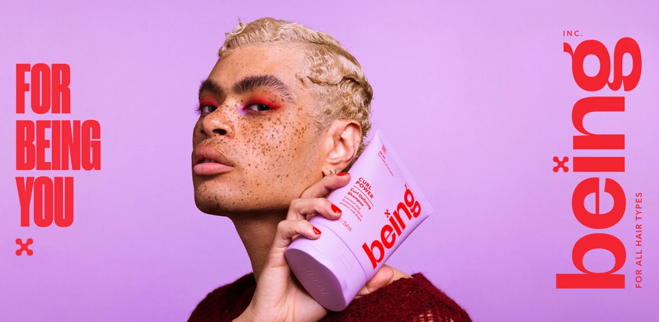 Close-up of a person with blonde wavy hair holding a tube of hair product in front of a light purple backdrop with red text.