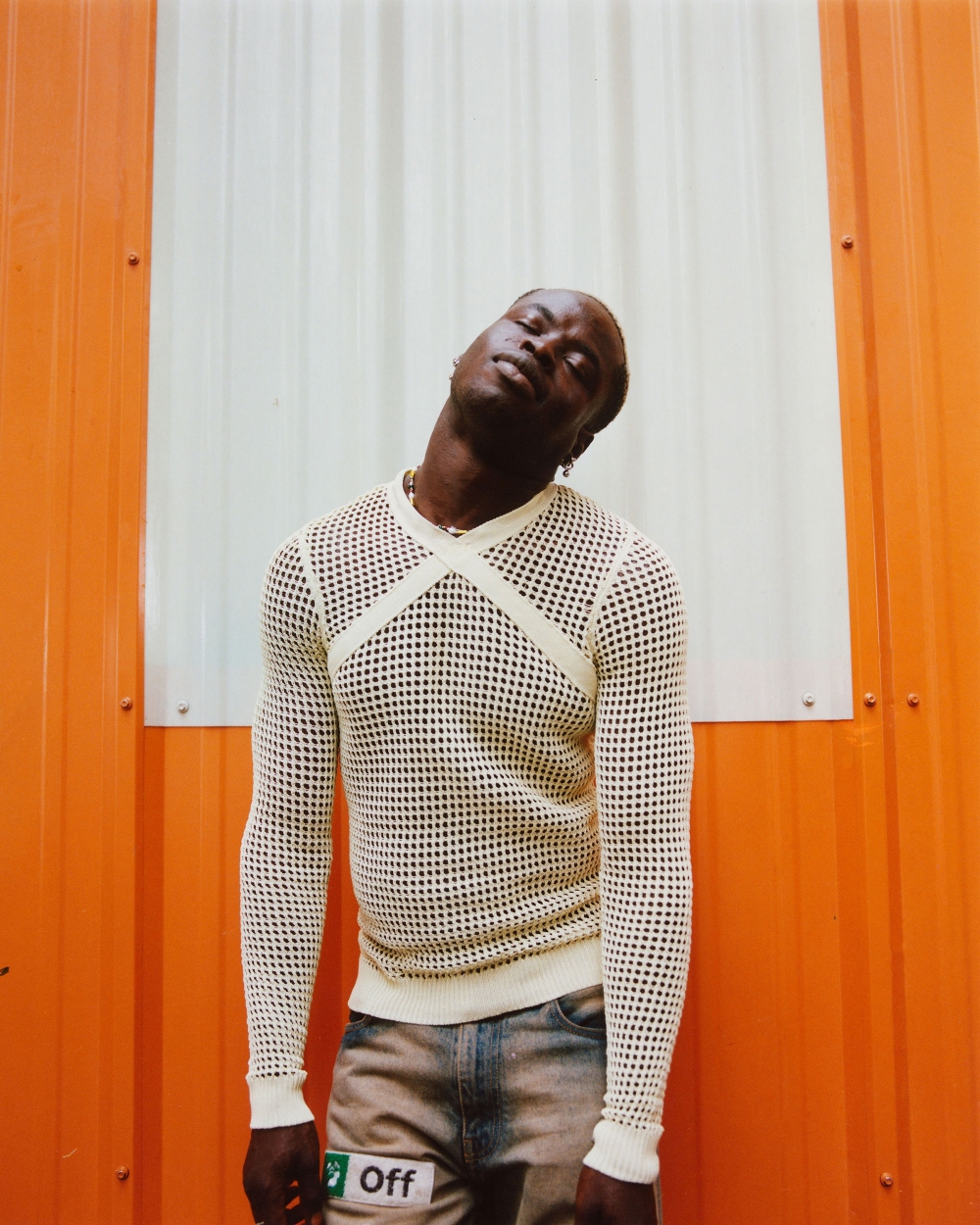 Eye-level shot of an adult man with dark skin wearing a white mesh long-sleeve shirt and distressed jeans against an orange and white wall.