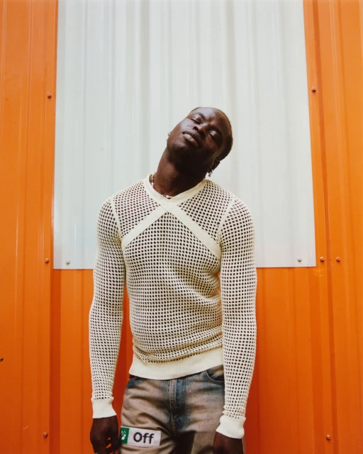 Eye-level shot of an adult man with dark skin wearing a white mesh long-sleeve shirt and distressed jeans against an orange and white wall.