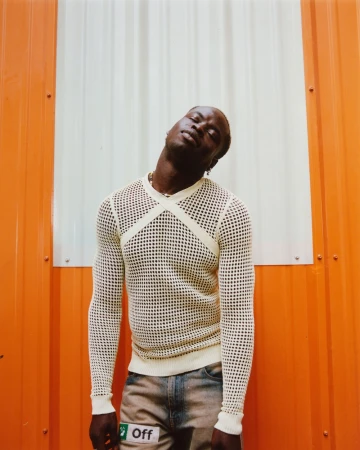 Eye-level shot of an adult man with dark skin wearing a white mesh long-sleeve shirt and distressed jeans against an orange and white wall.