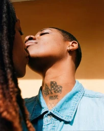 Close-up portrait of two people kissing outdoors in bright sunlight, wearing a blue denim shirt with a neck tattoo.