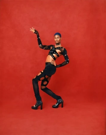 Black man with blue hair posing in black cutout top and trousers against a textured red background